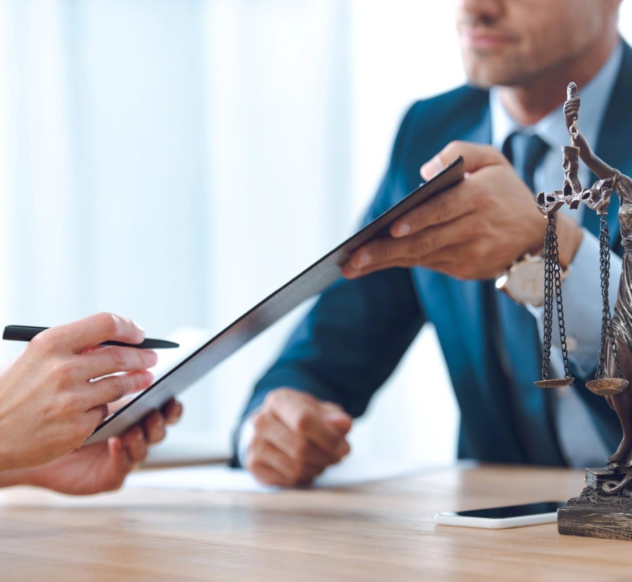 Two people in business attire exchanging a document at a desk, with a statuette of Lady Justice and a smartphone visible on the table.