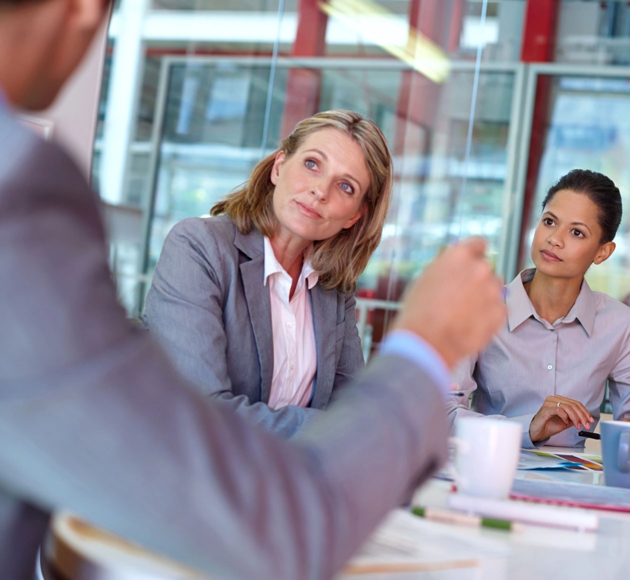 Two women in business attire sit at a conference table, listening to a man speak during a meeting in a modern office setting.