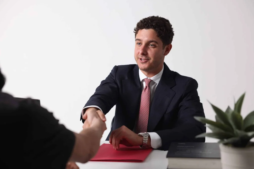 A man in a suit sits at a desk, smiling and shaking hands with another person. A red folder, books, and a potted plant are also on the desk, creating a welcoming home office atmosphere.
