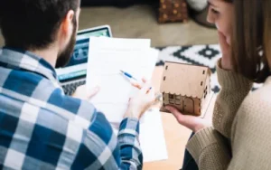 Two people sit together reviewing paperwork with a laptop; one holds a pen and the other holds a small model house, suggesting a discussion about home buying, real estate, or next steps after a car accident.