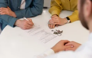 After a car accident, three people sit at a white table with documents and a set of keys; one person is signing papers while another observes.